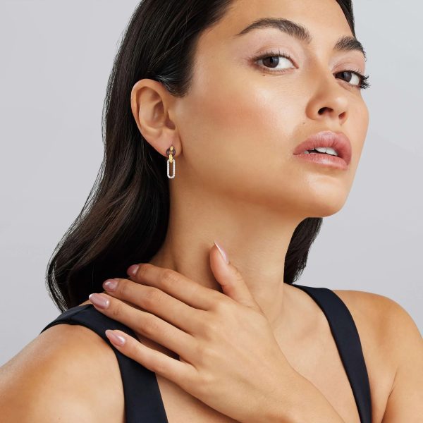 A woman with long dark hair, dressed in a black sleeveless top, gazes at the camera. She adorns her ears with LAGOS Signature Caviar Small 18K Gold and Black Ceramic Diamond Link Drop Earrings and sports a natural makeup look. Her right hand rests gently on her collarbone against a soft, neutral backdrop.