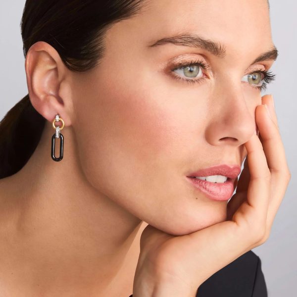 A woman with light skin and brown hair gazes thoughtfully into the distance. She is wearing a black top and adorned with LAGOS Signature Caviar 18K Gold and Black Ceramic Diamond Link Drop Earrings. Her hand rests against her cheek, and she has an expression of calm contemplation. The background is neutral.