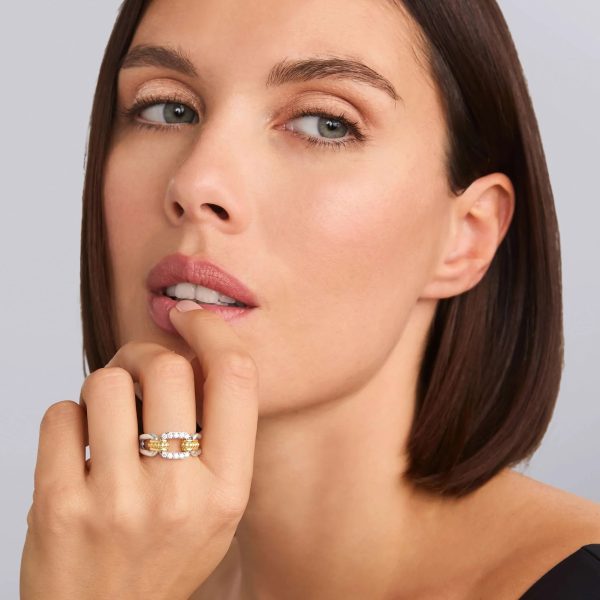 A woman with shoulder-length brown hair and minimal makeup is looking thoughtfully, holding her chin with her hand. She is displaying the LAGOS Signature Caviar Two-Tone Link Diamond Ring, featuring multiple bands and embedded stones. She is wearing a black top, and the background is a plain light gray.