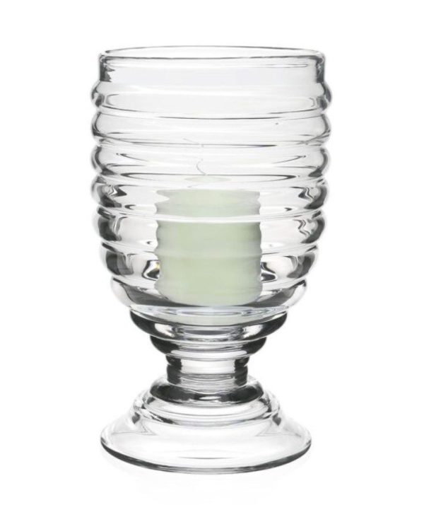 The William Yeoward Ripples Footed Hurricane - 11" is showcased against a white background. It features a ribbed design in clear glass, holding a small white candle within. The hurricane has a wide, sturdy base and its cylindrical shape gently flares out towards the top.