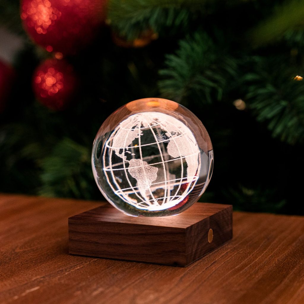 A glass globe rests on a wooden base, showcasing a detailed map of the world. Soft lighting highlights its intricate engravings. A blurred backdrop of festive decorations and greenery adds a warm, cozy atmosphere.