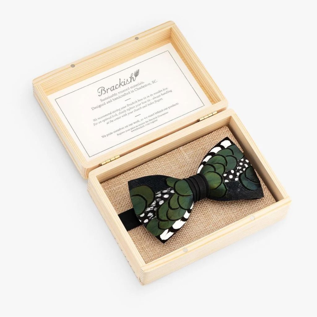 A wooden box with an open lid reveals a green and black bow tie with decorative patterns. The bow tie rests on a burlap-like fabric, and a card with text is attached inside the lid. The brand "Brackish" is printed on the card.