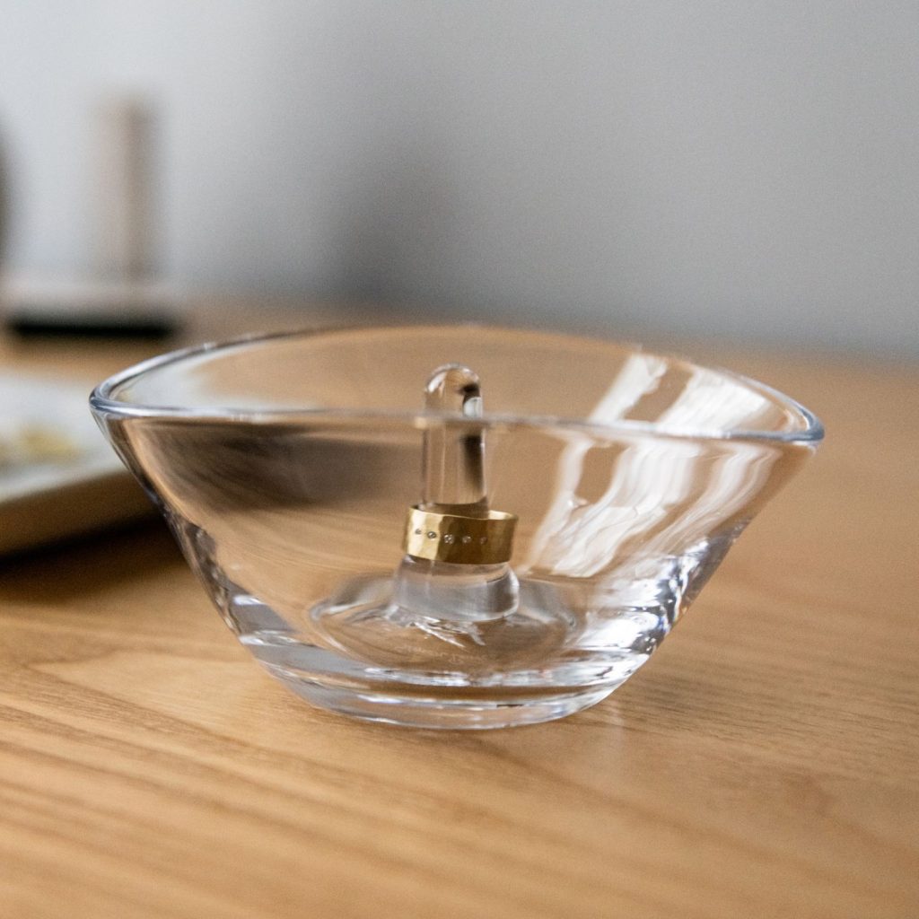 A small, transparent glass bowl sits on a wooden surface, containing a ring-shaped object with a vertical element protruding from the center. The background is blurred, hinting at a calm indoor setting.