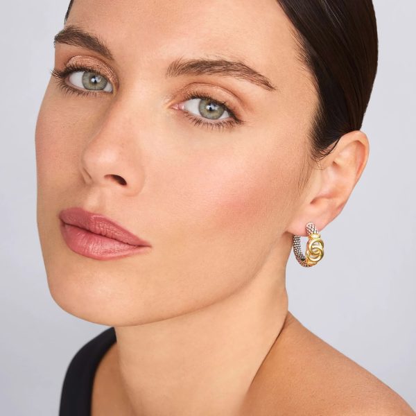 Close-up of a person with light skin, short dark hair, and light hazel eyes. They are wearing LAGOS Signature Caviar Two-Tone Interlocking Hoop Earrings and a black, off-shoulder top. The background is plain and light-colored, highlighting their minimalist style and elegant features.