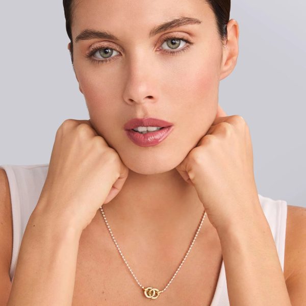 A woman with fair skin and short brown hair poses against a neutral background. She is wearing the LAGOS Signature Caviar Two-Tone Interlocking Pendant Necklace, beautifully showcasing its delicate design with two intertwined circles. Her elbows are raised, and her hands gently touch her chin, drawing attention to the elegant necklace.