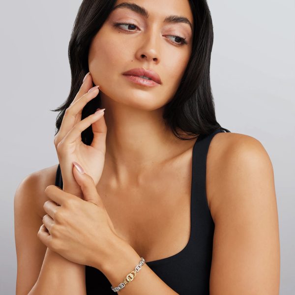 A woman with long dark hair is gazing to the side while gently touching her face with one hand. She is wearing a sleeveless black top and showcasing the LAGOS Signature Caviar Two-Tone Interlocking Diamond Link Bracelet on her wrist. The background is a smooth gradient.