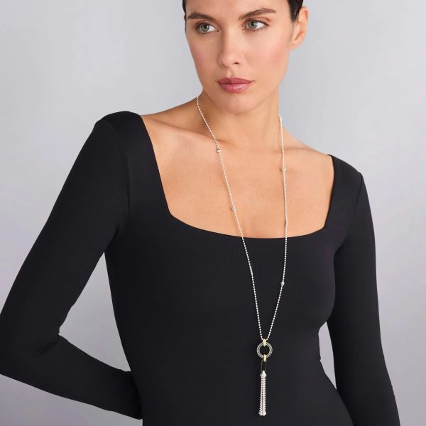 A woman with brown hair tied back wears a fitted, long-sleeve black dress with a wide, square neckline. She accessorizes with the LAGOS Black Caviar Two-Tone Black Ceramic Tassel Pendant Necklace, featuring an elegant circular pendant and tassel, looking off to the side against a neutral background.