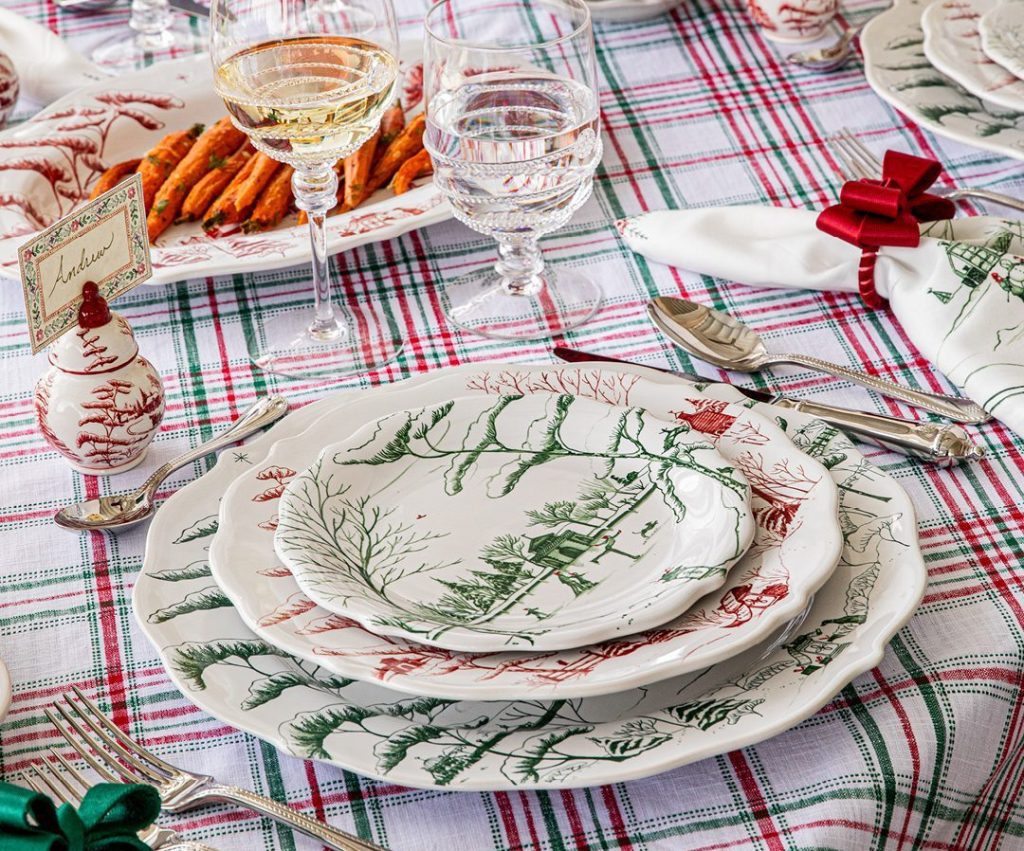 Festive table setting featuring holiday-themed plates on a red and green plaid tablecloth. Includes wine glasses, silverware, and a small menu card. Napkins are tied with festive bows.