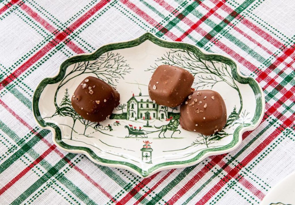 A festive table setting featuring a decorative plate with a winter scene, holding four chocolate truffles sprinkled with sea salt. The tablecloth has a red and green plaid pattern, creating a holiday atmosphere.