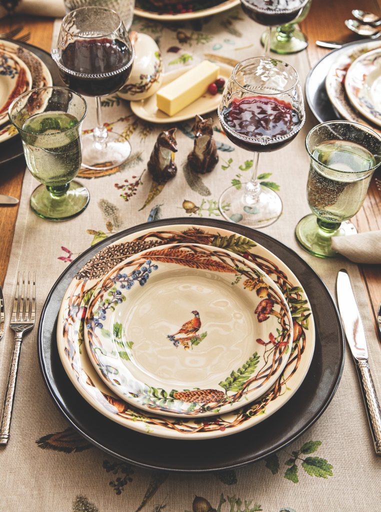 A beautifully set dining table features vintage-style plates with bird designs, elegant silverware, and glasses of red wine and sparkling water. A linen table runner with floral patterns complements the rustic theme.