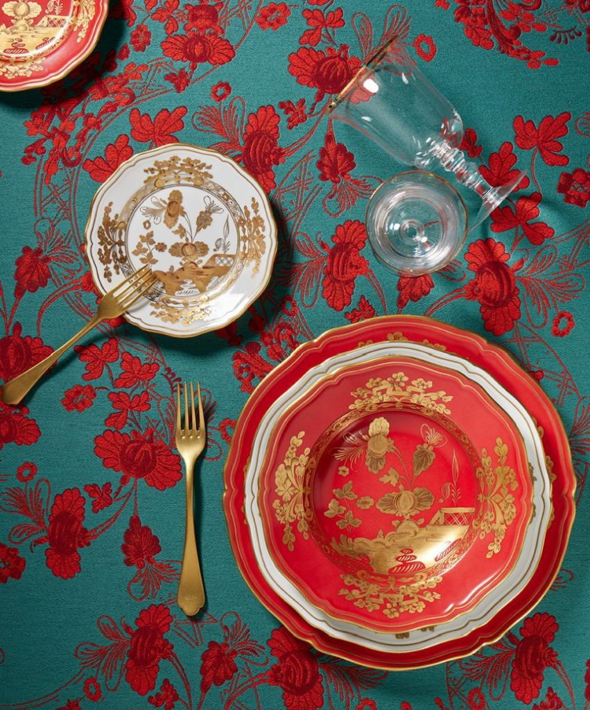 A red and gold floral-patterned dinner set lies on a vibrant teal tablecloth with red floral designs. The setup includes plates, a glass, gold cutlery, and a detailed decorative theme, creating an elegant dining scene.