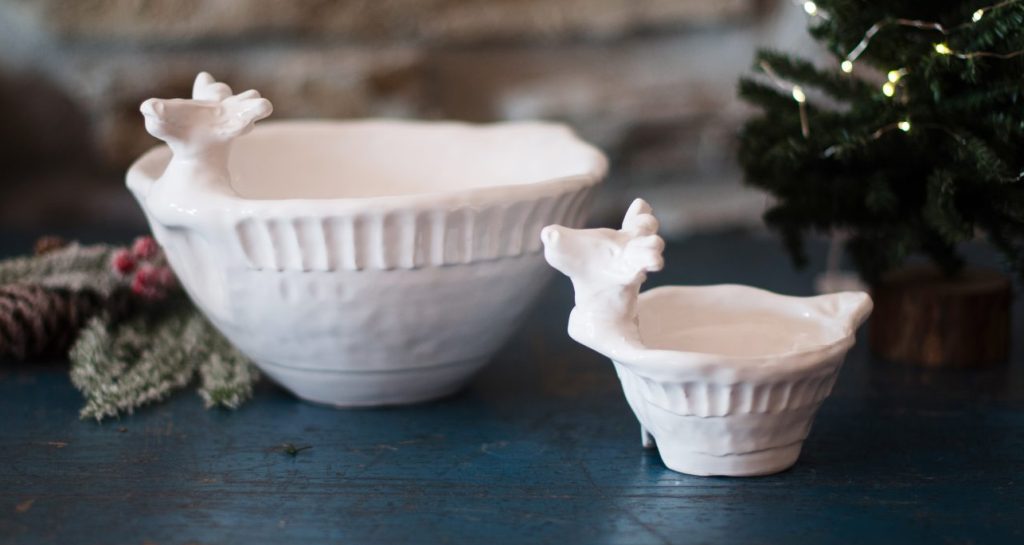 Two white ceramic bowls with deer head handles are placed on a blue wooden surface. They are surrounded by small, decorated faux Christmas trees and pinecones, against a stone wall backdrop.