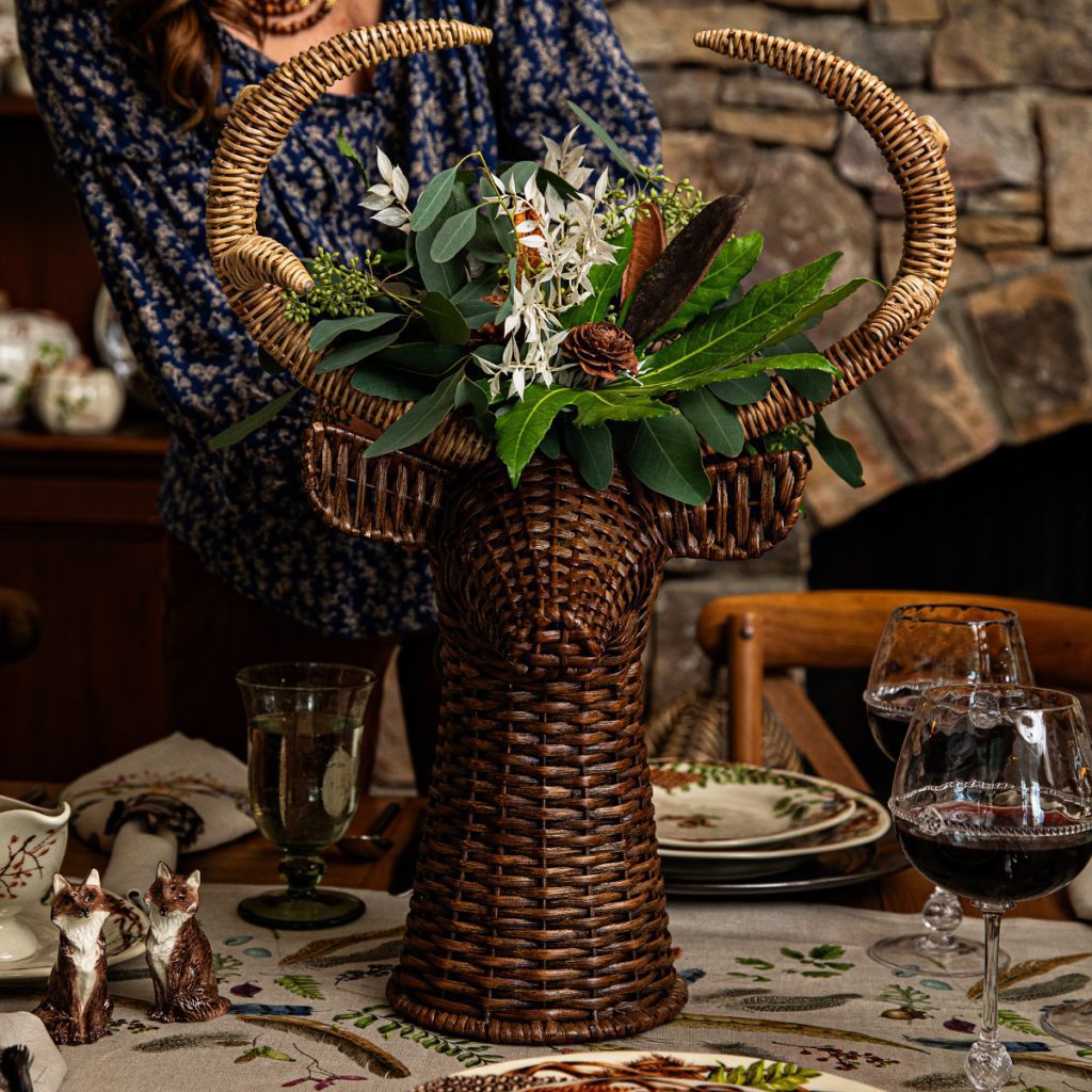 A wicker bull-shaped vase filled with green foliage and white flowers is placed on a dining table set with dishes and glasses. A person in a blue patterned shirt stands behind it. A stone wall and decorative items are in the background.