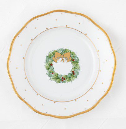 The Herend Christmas Wreath Dessert Plate is a decorative porcelain piece with a gold rim, showcasing a festive design featuring a green holly wreath adorned with red berries, centered around a bright golden bow.