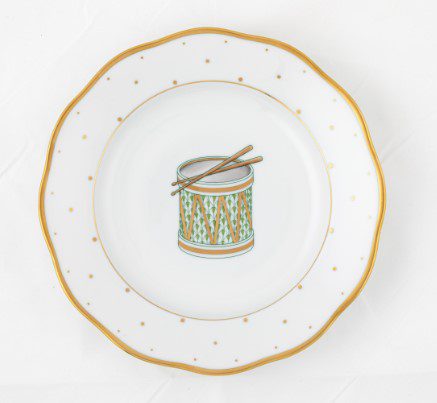 The Herend Christmas Drum Dessert Plate is a decorative white plate adorned with a golden edge and small dots, featuring an illustration of a drum with two crossed drumsticks at its center.