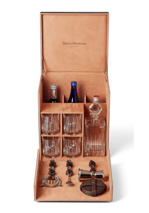 The Reed and Barton 200th Anniversary Leather Portable Bar is a luxurious bar set housed in an open wooden box, showcasing glass decanters, bottles, crystal glasses, and essential bar tools like a corkscrew and jigger. The interior is elegantly lined with peach-colored fabric.