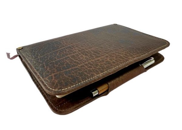 The Kingfisher Leatherworks Bison Leather Portfolio features a textured leather notebook cover with a pen holder, including both a notebook and a pen. The slightly worn brown leather offers it an appealing vintage look. The cover is closed and laid flat.