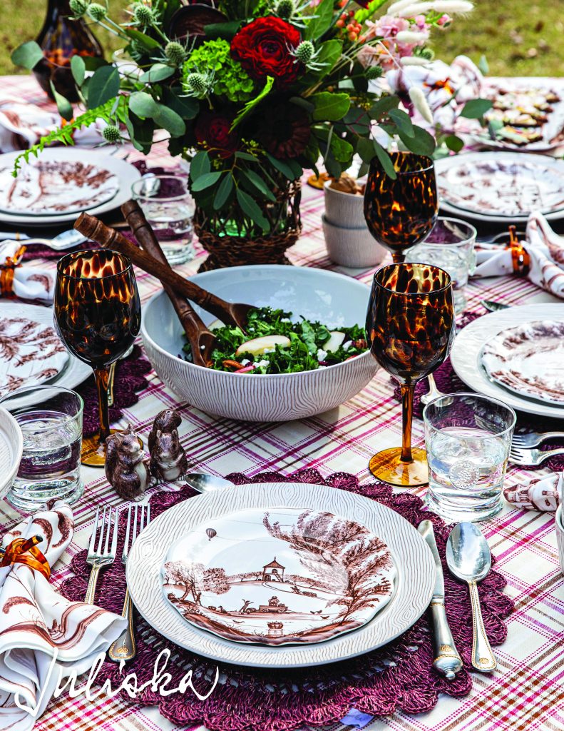 A beautifully set outdoor table with patterned plates, amber glasses, a white bowl with salad, and decorative napkins. The centerpiece is a floral arrangement with red roses and greenery, all on a plaid tablecloth.