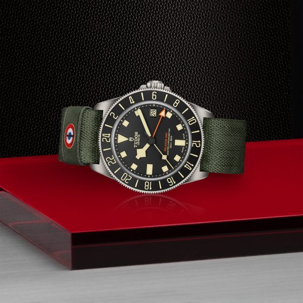 A luxury wristwatch with a green fabric strap and black dial set on a red and black display stand. The watch features luminous markers and hands, with a date window and a black bezel.