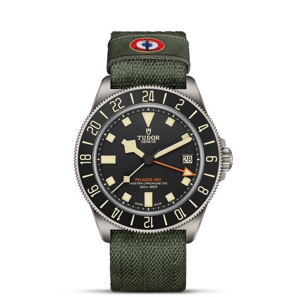 A Tudor Pelagos GMT watch with a green fabric strap and black dial. The watch displays the date and has luminous hands and markers. The bezel is marked with a 24-hour scale, and the strap has a circular logo patch.