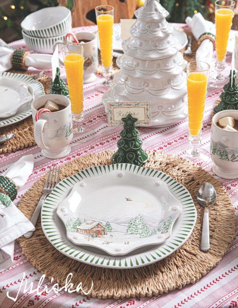 A festive table setting with white and green holiday-themed plates showcasing a winter scene. There are orange juice glasses, decorative trees, a white ceramic Christmas tree centerpiece, and mugs with candy canes on a striped tablecloth.