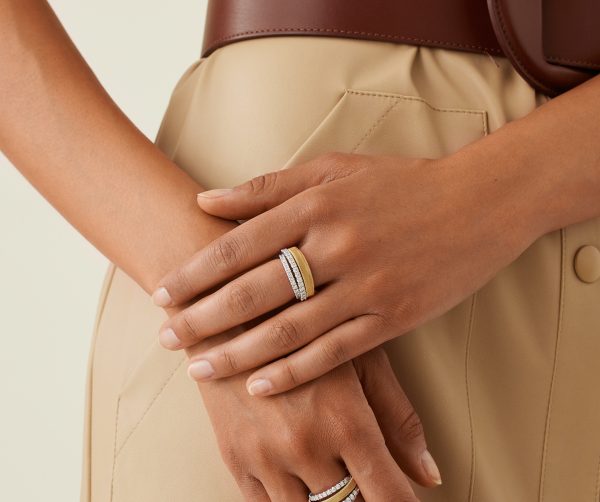 A person in a beige outfit with a brown belt is clasping their hands, showcasing a striking Marco Bicego Yellow Gold 4-Strand Coil Ring embellished with pavé diamonds among other rings. The emphasis is on the hands and the elegant rings.