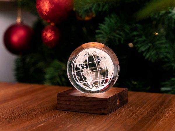 The Ginko Design Crystal World Paperweight on Wood Light Box, featuring a precisely etched world map, sits beautifully on a table. It emits a warm glow against blurred red baubles and green foliage in the background, enhancing the festive ambiance.