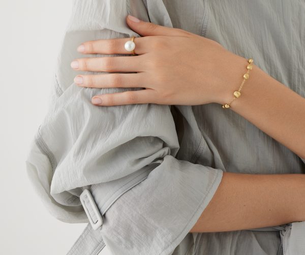 A close-up of a person wearing a sheer, light gray blouse with their left arm across their chest, adorned with the Marco Bicego Africa Yellow Gold Station Bead Bracelet. They also have a ring with a large white pearl on their ring finger.