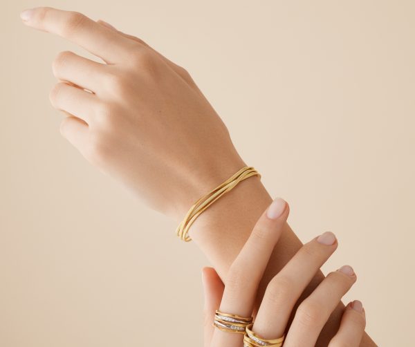 A close-up shot showcases hands adorned with gold rings and the Marco Bicego Marrakech 3 Strand Coil Bracelet. The hands are elegantly posed against a soft beige background, highlighting the jewelry.