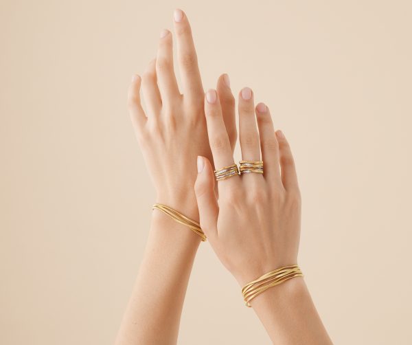 The image features two hands elegantly posed against a neutral background, adorned with exquisite gold jewelry. Among the adornments is the Marco Bicego Marrakech 3 Strand Coil Bracelet gracing the wrist, along with delicate rings on the fingers. The nails are beautifully manicured with a soft, light pink polish.