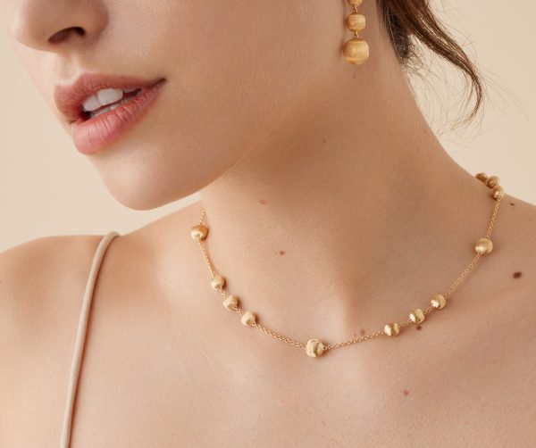 A close-up shot features a person adorned with the Marco Bicego Yellow Gold Station Bead Necklace, showcasing its exquisite bead-like details. They are also wearing complementary gold earrings. The backdrop is a neutral beige, and the person wears a thin strap top.