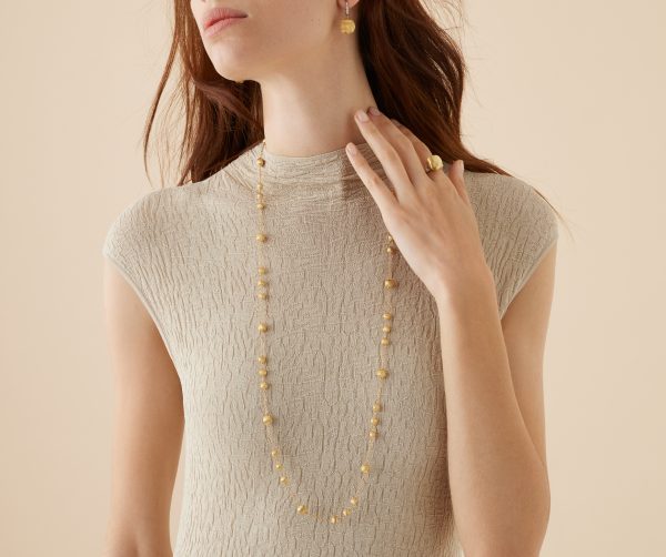 A woman with long brown hair is wearing a textured beige top, accessorized with the Marco Bicego Africa Yellow Gold Station Bead Long Necklace, matching earrings, and a gold ring. She has her left hand near her collarbone against a neutral background.