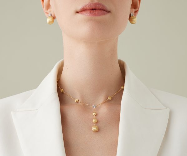 A close-up of a woman wearing a white blazer. She is adorned with gold pearl earrings and the Marco Bicego Yellow Gold Lariat Necklace With Diamonds, featuring a dangling pearl design. The background is a soft beige.