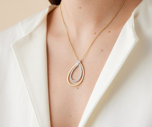 A person wearing a white blouse is adorned with the Marco Bicego Masai Yellow Gold Pendant Necklace, which features a gold teardrop shape with diamonds and an elegant layered design.