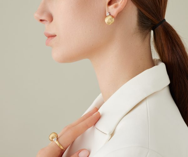 A woman with a ponytail wears a white blazer, showcasing the Marco Bicego Africa Yellow Gold Stud Earrings With Diamonds and a matching gold ring with gemstones. Her hand lightly touches her collar against a soft, neutral background.