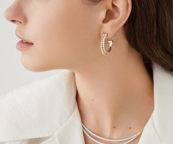 The woman is adorned with a pair of Marco Bicego Masai Yellow Gold Double Hoop Earrings with Diamonds, paired with a matching necklace. She complements her look with a white textured blazer, and her hair is neatly tucked behind her ear against a neutral light-colored background.