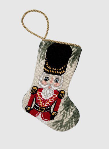 A festive stocking featuring a needlepoint design of a nutcracker soldier in red and gold attire. It has a loop for hanging and a background that appears to have green foliage.