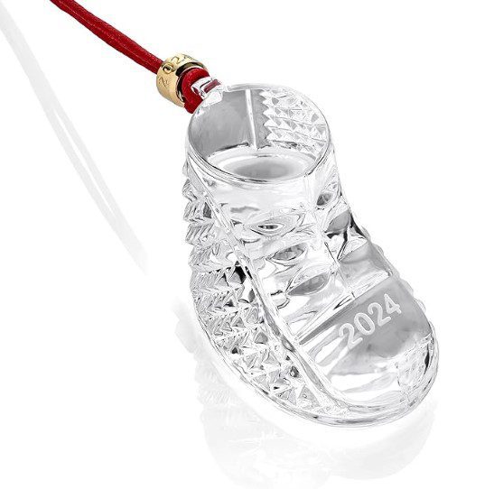 The Waterford 2024 Baby's First Christmas Ornament is a crystal baby shoe adorned with a red cord and gold accents, featuring decorative patterns and "2024" etched on the sole.