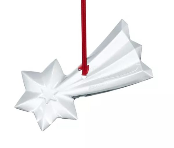 The Baccarat 2024 Noel Shooting Star Ornament is a white ceramic star with a red ribbon, featuring a glossy finish and an angular design reminiscent of a shooting star.