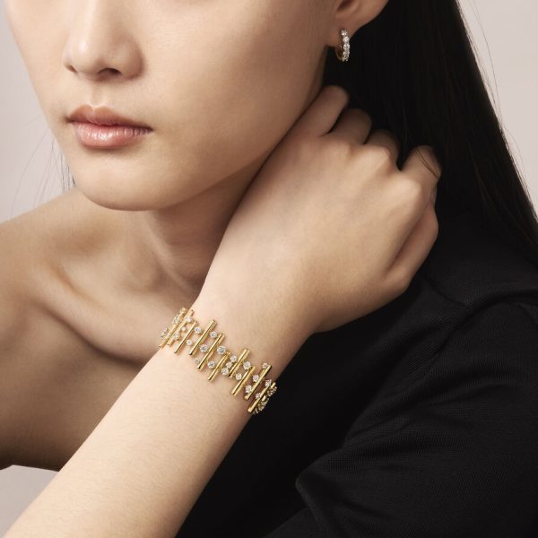 A person is wearing the Hearts on Fire Barre Multi-Row Bracelet, which features diamond accents, on their wrist and a pair of small gold hoop earrings adorned with diamonds. The individual has long dark hair and is dressed in black.