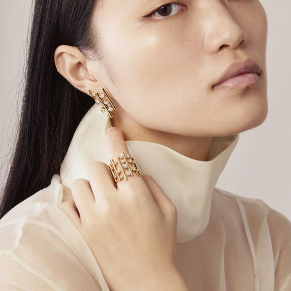 A woman adorned with a Hearts on Fire Barre Multi-Row Diamond Ring, complemented by gold earrings that feature delicate pearl accents, gently touches her earlobe. Her long black hair cascades over a sheer, light-colored turtleneck top against a neutral background.