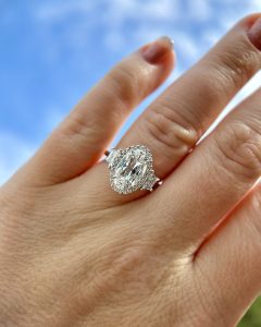 A beautiful trendy engagement ring with diamonds rests on a woman’s hand against a blue sky with clouds.