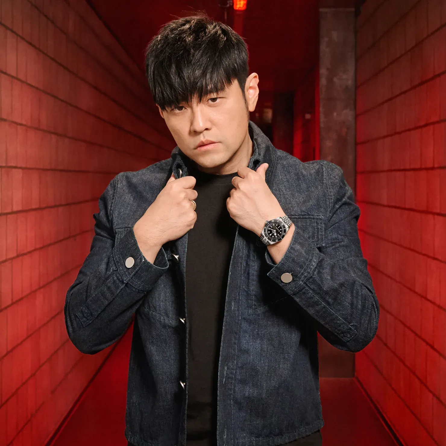 TUDOR Ambassador Jay Chou posing in a red hallway wearing a dark jacket and TUDOR watch