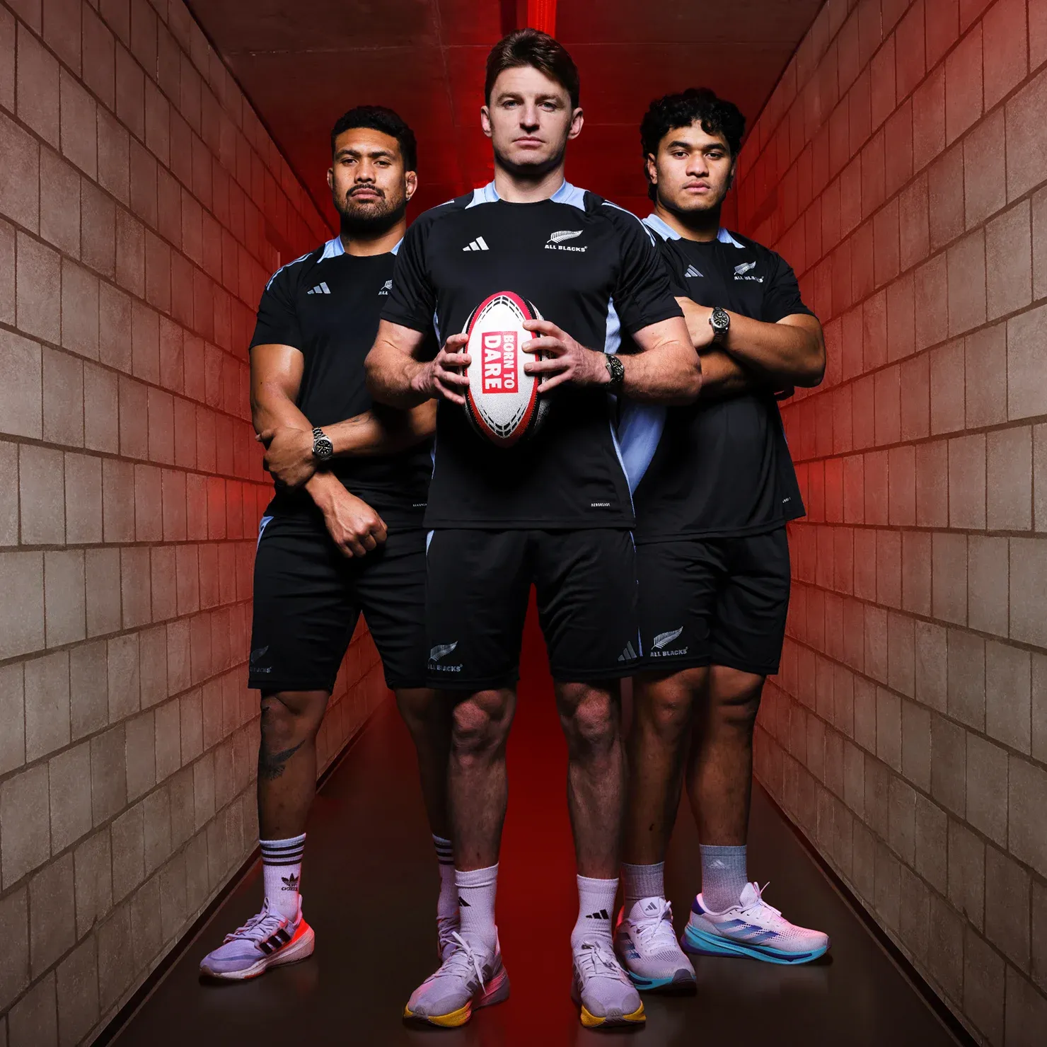 All Blacks rugby players in hall with brick wall and red lighting holding rugby ball