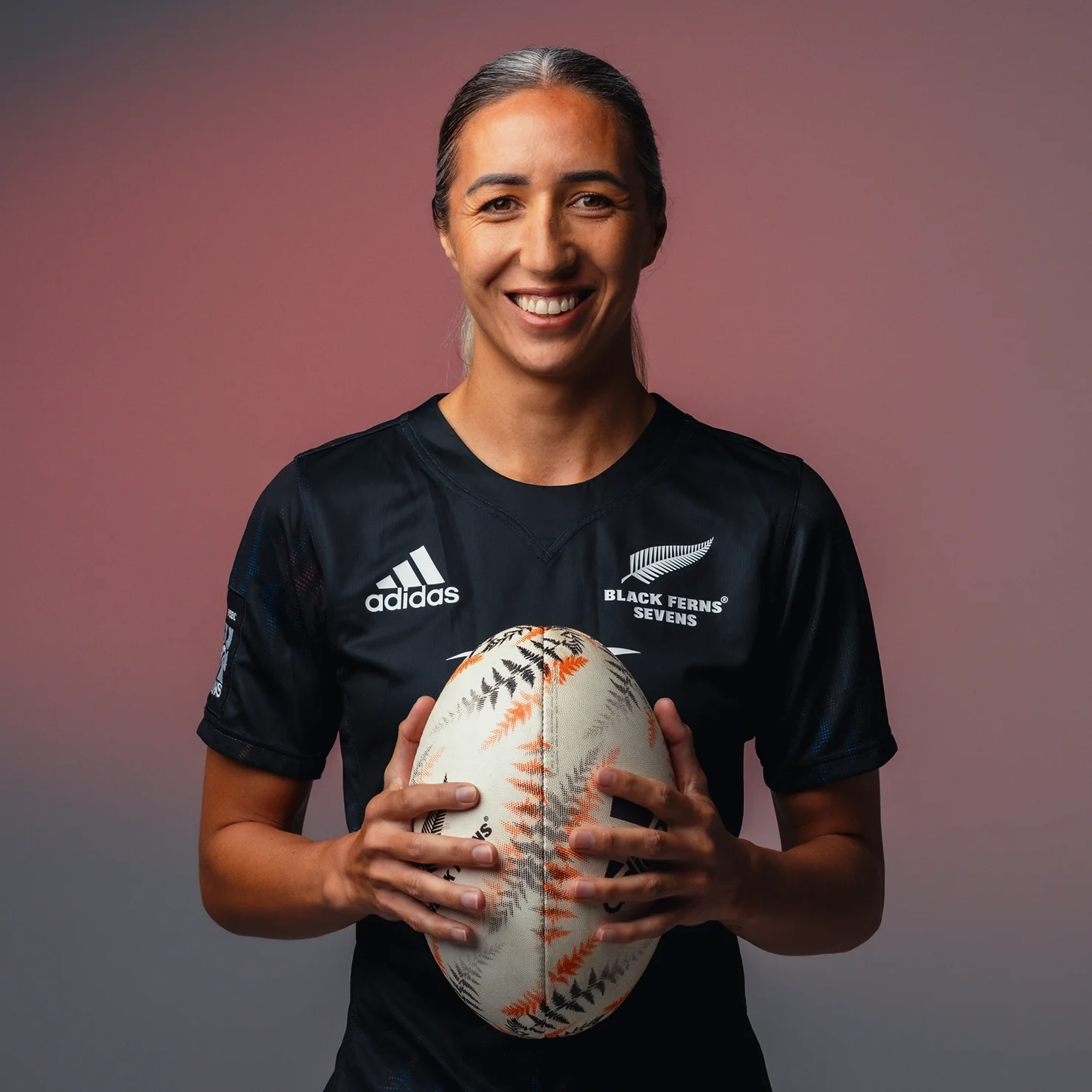 TUDOR Ambassador Sarah Hirini wearing a black rugby jersey and smiling while holding a rugby ball against a neutral background