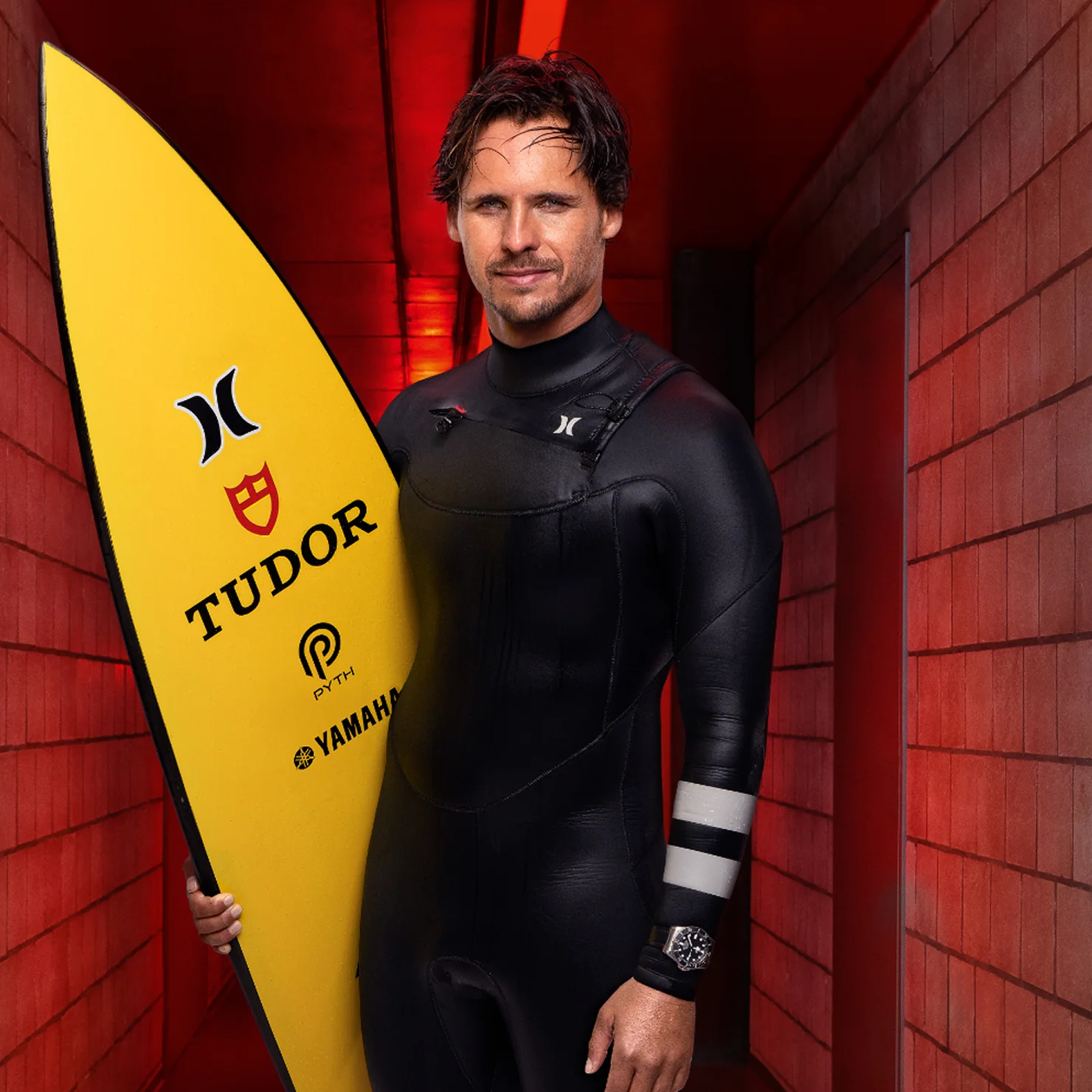 TUDOR Ambassador Nic von Rupp in a black wetsuit holding a yellow surfboard with TUDOR and Yamaha logos, standing in a red tunnel