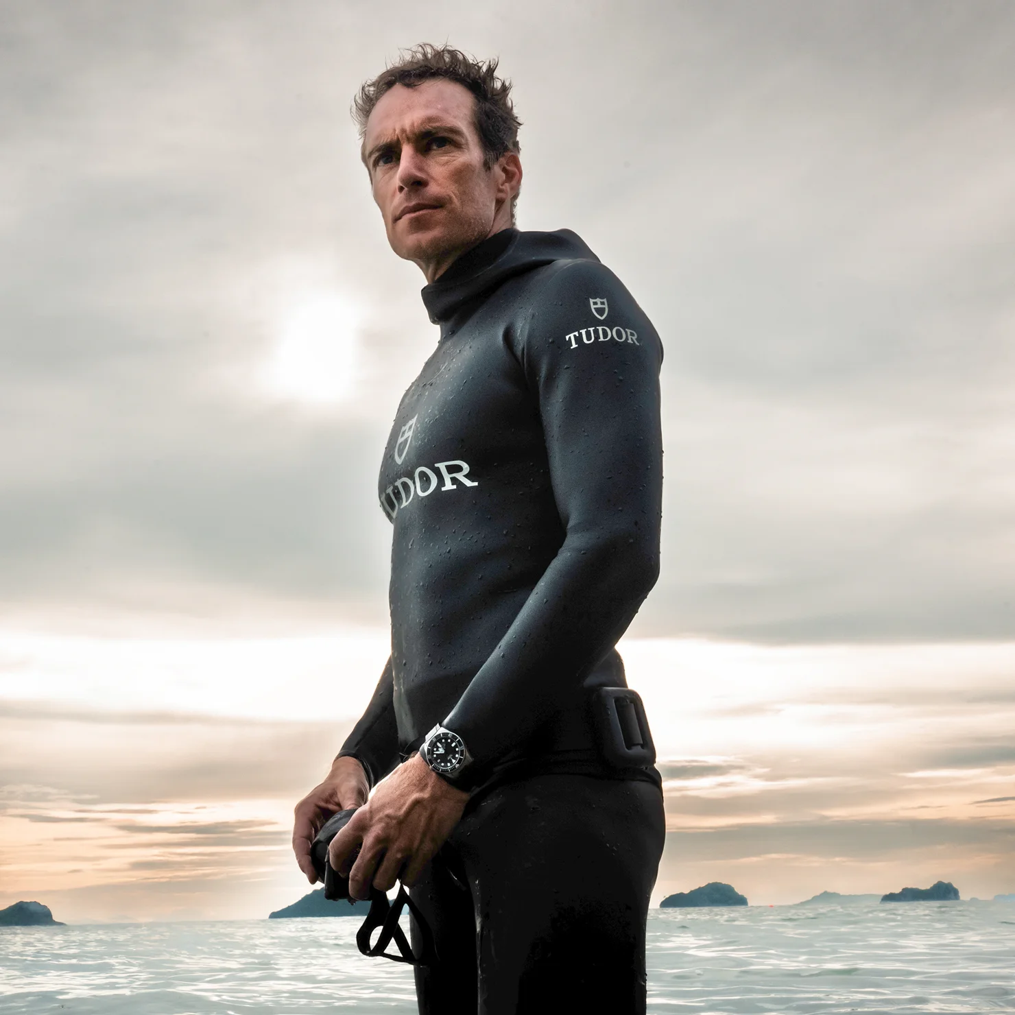 TUDOR Ambassador Morgan Bourc’his in a black wetsuit standing confidently by the ocean with islands in the background, showcasing a TUDOR watch on his wrist.