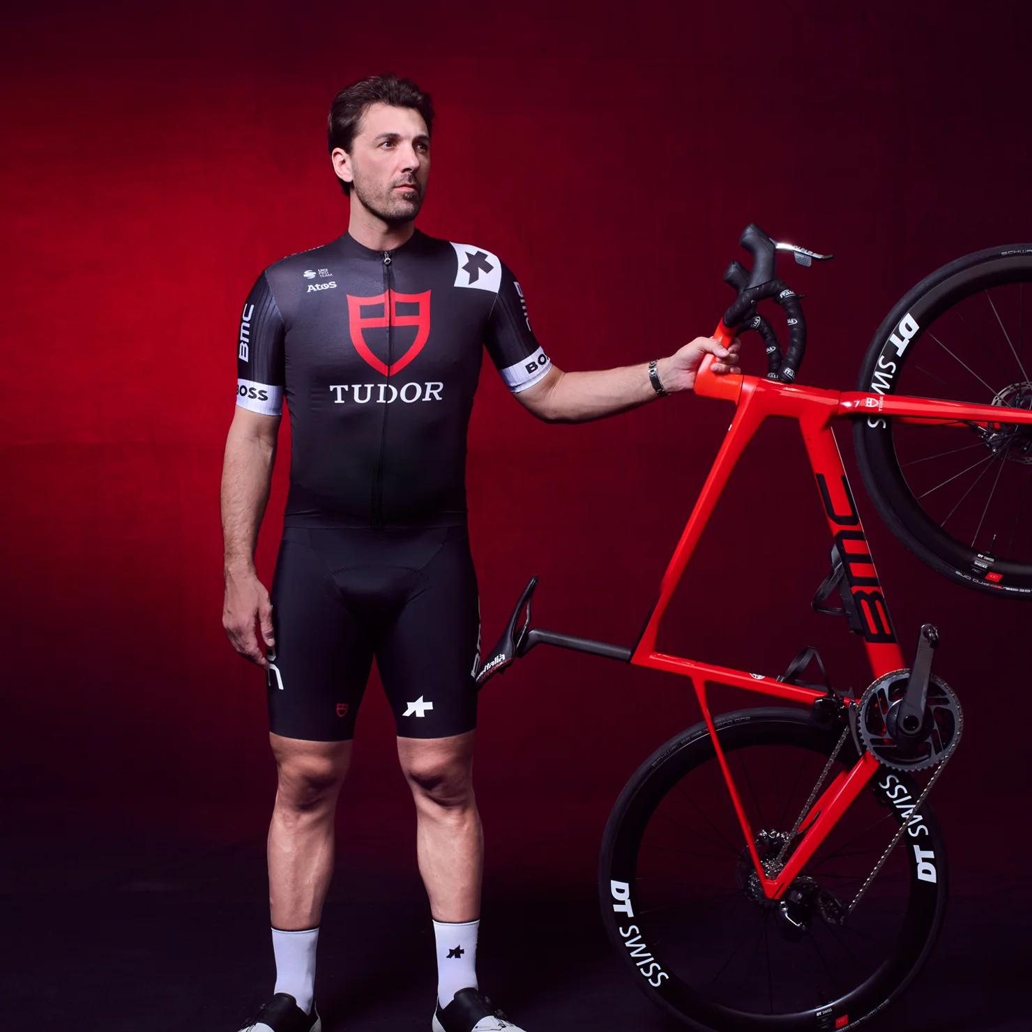 TUDOR Ambassador Fabian Cancellara wearing a black and red cycling kit, standing beside a red road bike against a dramatic red-lit background