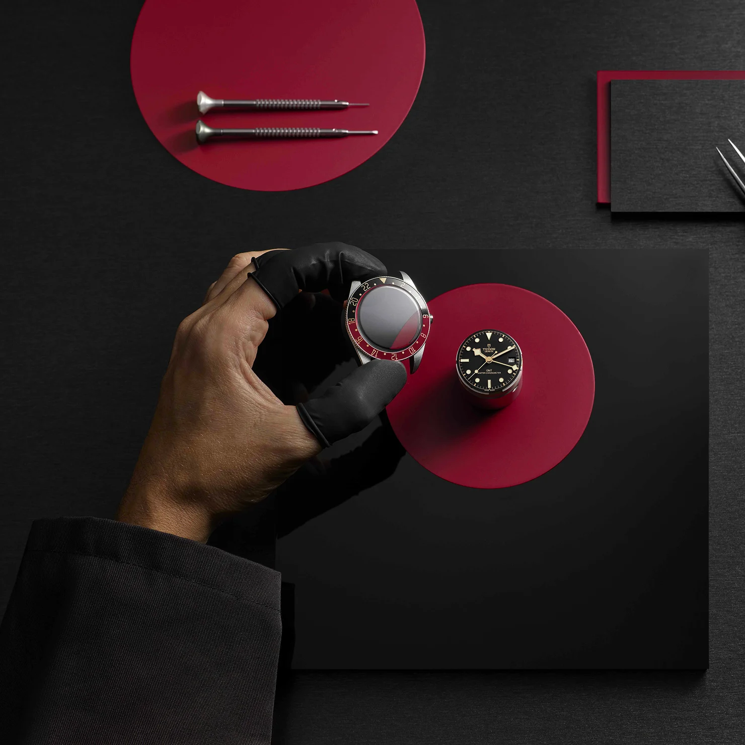 Watchmaker's gloved hand holding a partially assembled TUDOR watch case above a watch dial